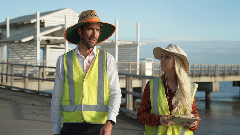 Two workers wearing high-vis vests, discussing near waterfront pier, Australia.
